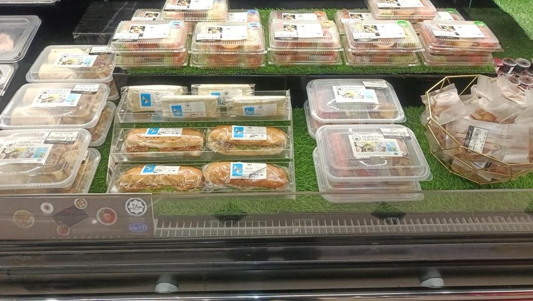 A store chiller stacked with sealed Foodplus packs: salad trays, wrapped sandwiches and chicken buns, with the Halal and MeSTI stickers on the glass